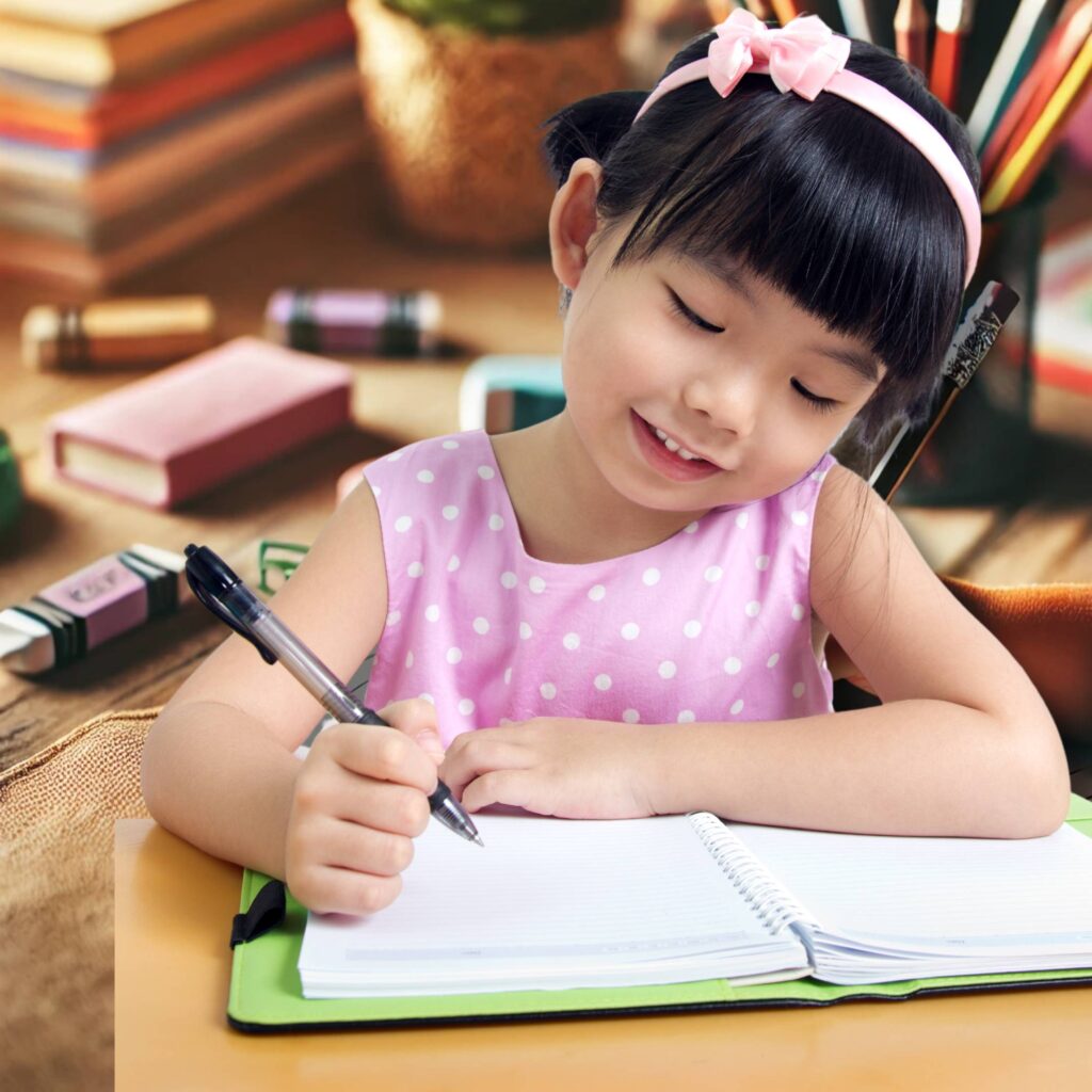 A child's hand writing neatly in a notebook surrounded by colorful stationery.