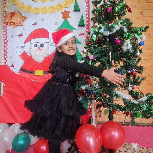 A smiling child in a black dress and Santa hat posing joyfully by a Christmas tree decorated with ornaments, surrounded by festive balloons and gifts in a preschool setting.