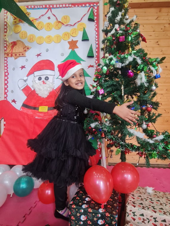 A smiling child in a black dress and Santa hat posing joyfully by a Christmas tree decorated with ornaments, surrounded by festive balloons and gifts in a preschool setting.