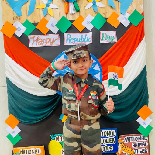 A young Udayan Kidz preschool student dressed as a soldier, saluting with a patriotic backdrop featuring the Indian flag and national symbols.